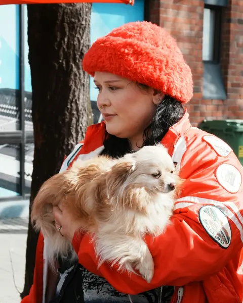 Una mujer con abrigo rojo lleva en brazos a su perro pequeño